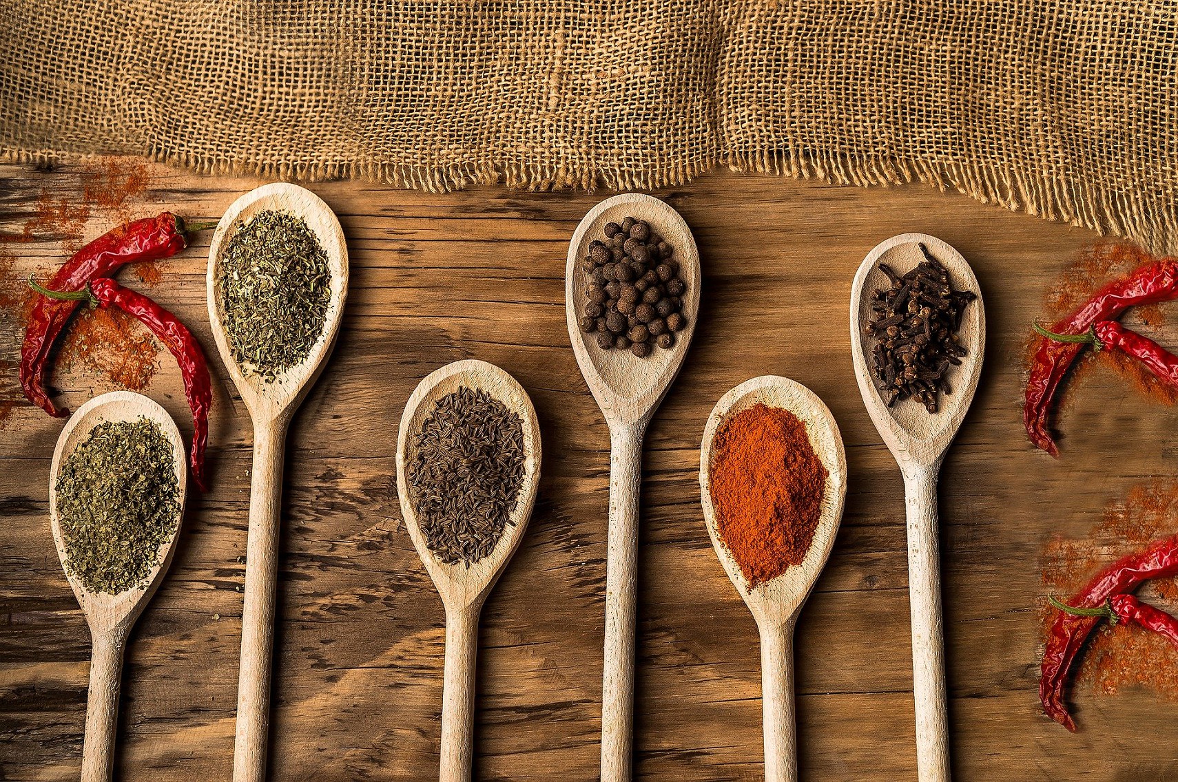 A series of wooden spoons, each holding a different spice, on a textured wooden background
